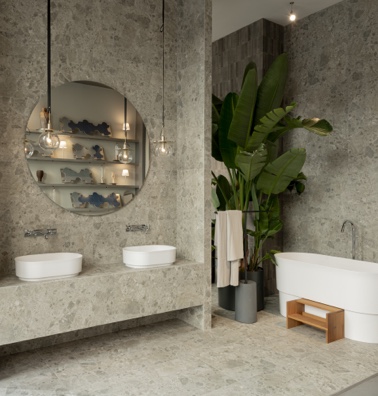 Modernes Badezimmer aus Natursteinfliesen mit Badewanne, zwei Waschbecken und einem großen, runden Spiegel | Bauzentrum Zillinger